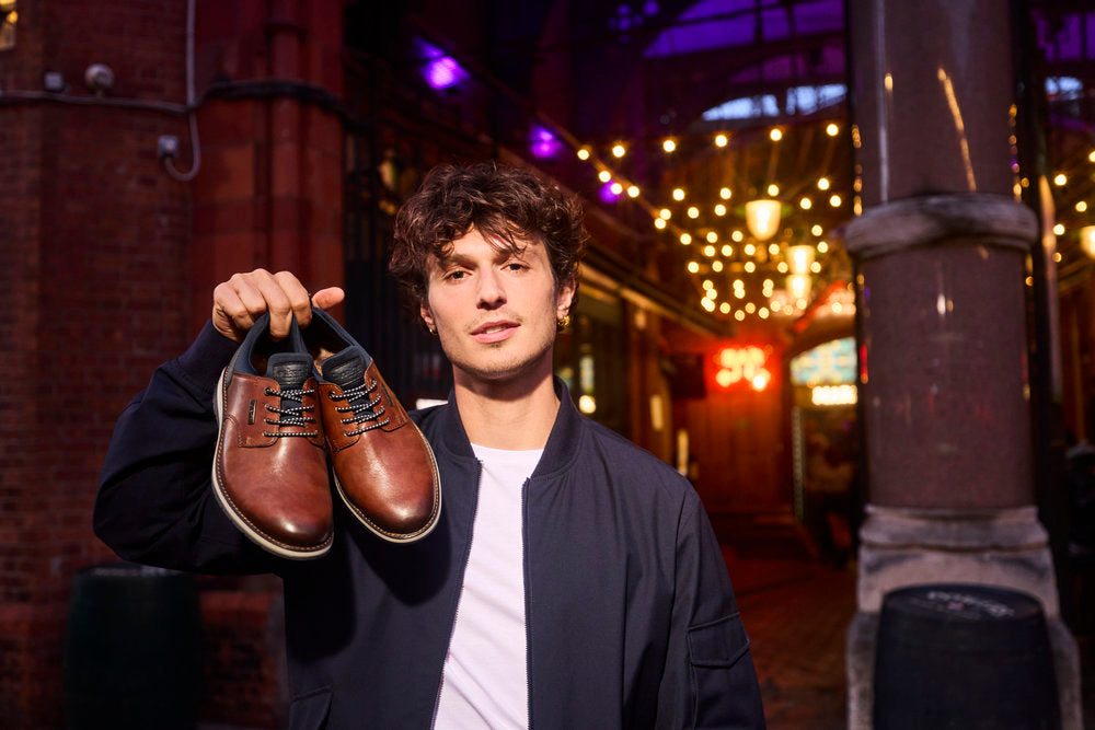 A man holding up a pair of brown leather lace-up dress shoes, wearing a dark navy bomber jacket over a white t-shirt, standing in a dimly lit urban alleyway at night with warm string lights in the background.